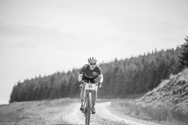 Monochrome image of a male mtb cyclist, number 40, intensely riding downhill on a gravel trail with dense pine forests on either side, in a focused racing posture