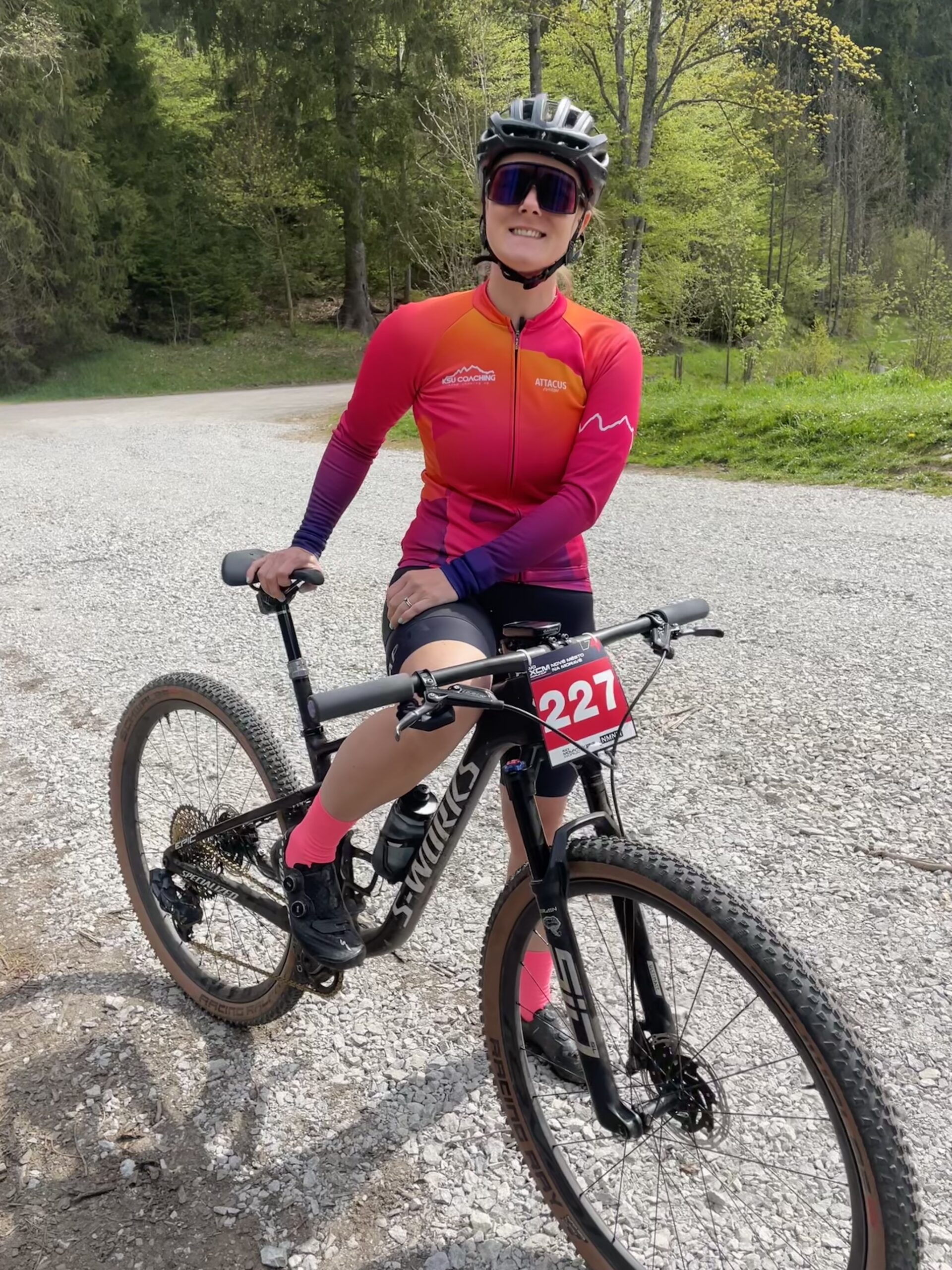 Smiling female mtb cyclist in a pink and orange jersey posing with her mountain bike, number 227, on a gravel path surrounded by lush green forest