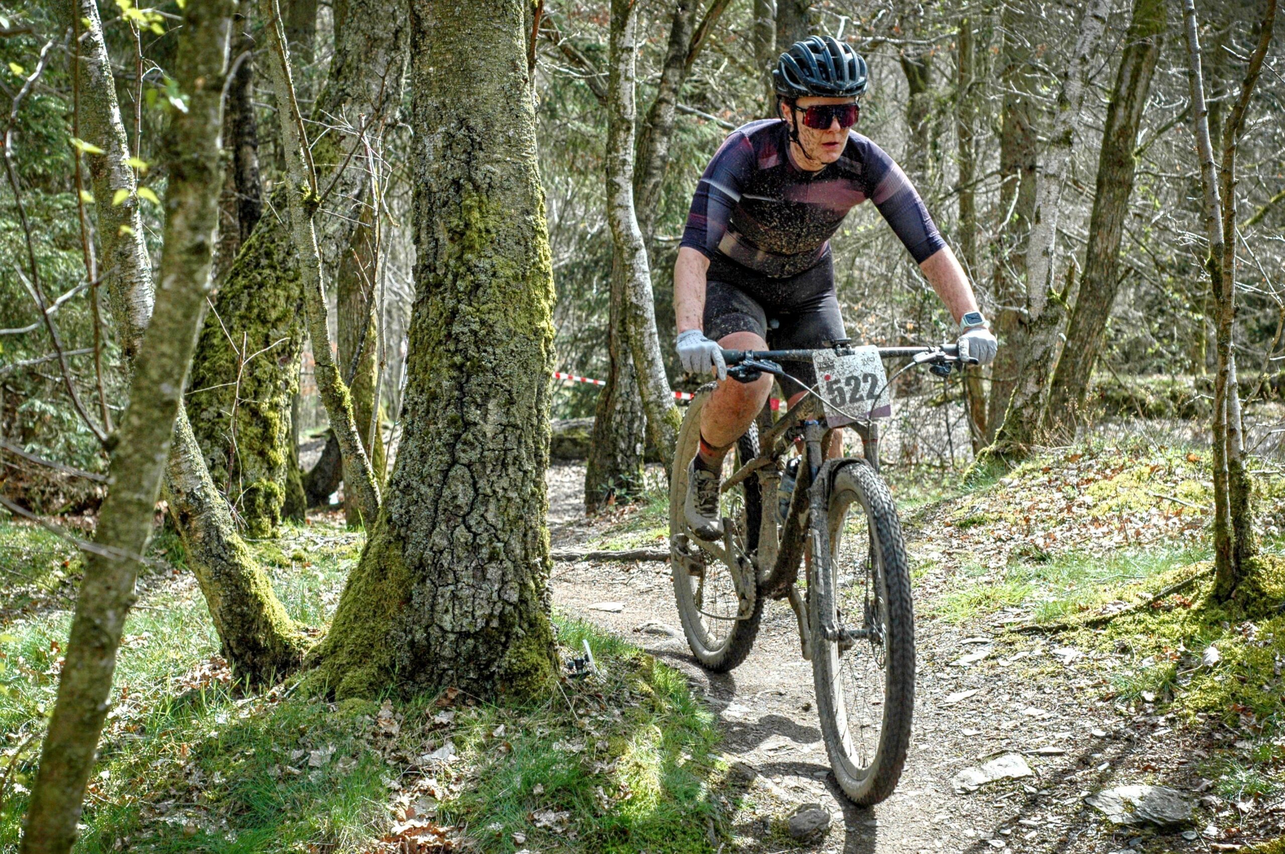 Mountain biker riding through a forest trail, wearing a helmet, sunglasses, and cycling gear, with a race number 522 on the bike