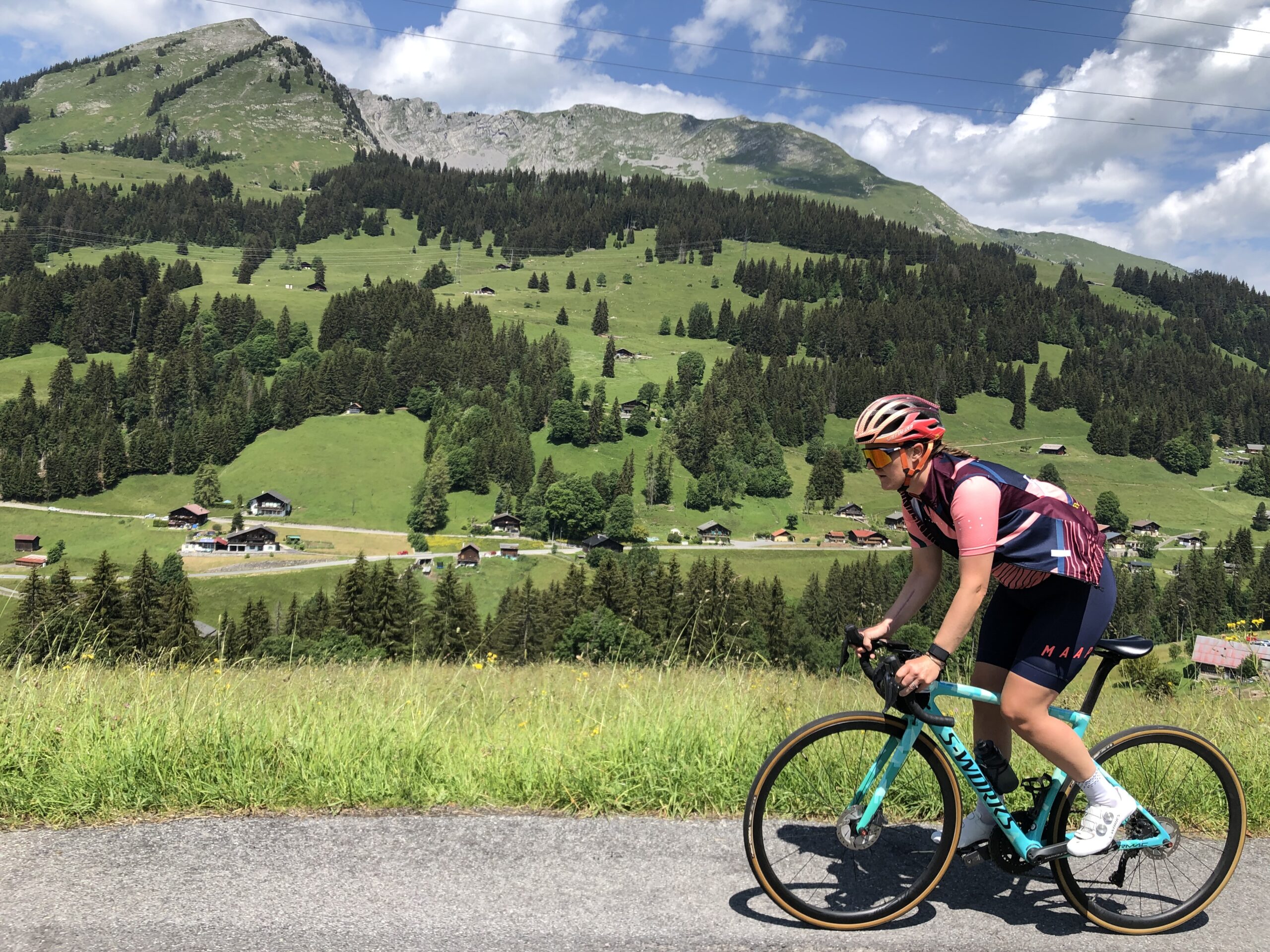 Female cyclist in a pink and navy kit riding a turquoise road bike on a scenic mountain road, with lush green hills and charming alpine cottages in the background under a bright blue sky.