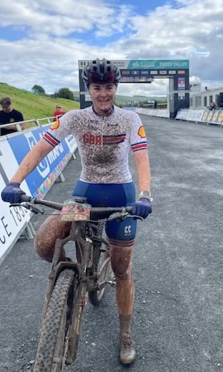 Mountain biker standing with their bike at the finish line, covered in mud, wearing a GBR jersey, helmet, and gloves, smiling after completing a race.