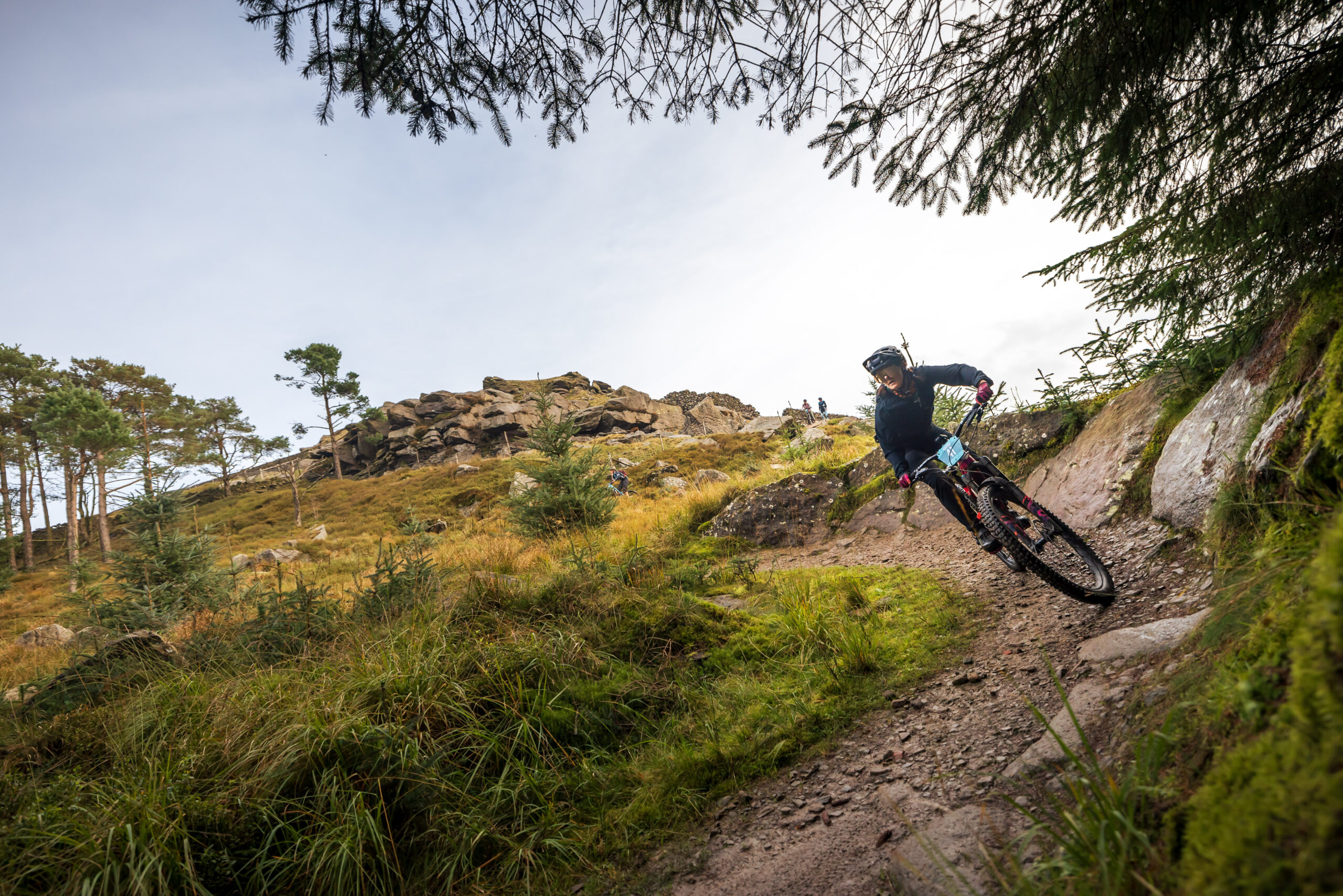 A mountain biker wearing protective gear, including a helmet and sunglasses, rides downhill on a rugged, rocky trail. The rider leans into a curve, navigating the challenging terrain surrounded by tall grass and scattered rocks. In the background, the landscape features rocky outcrops, sparse trees, and a clear sky, indicating an outdoor mountain biking scene in a natural setting. The photo captures the dynamic motion and intensity of the sport