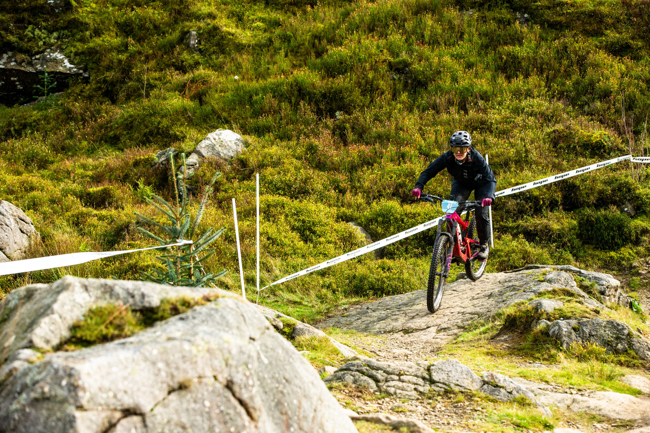 A mountain biker wearing a helmet and protective gear rides downhill on a rocky trail. The rider is approaching a technical section of the course, with rocks and uneven terrain visible under the bike. The trail is lined with white tape, marking the course boundaries, and the surrounding landscape is covered in green vegetation and small trees. The rider maintains a focused and controlled posture, ready to navigate the challenging descent. The image captures the intensity and concentration required for mountain biking on rugged terrain.