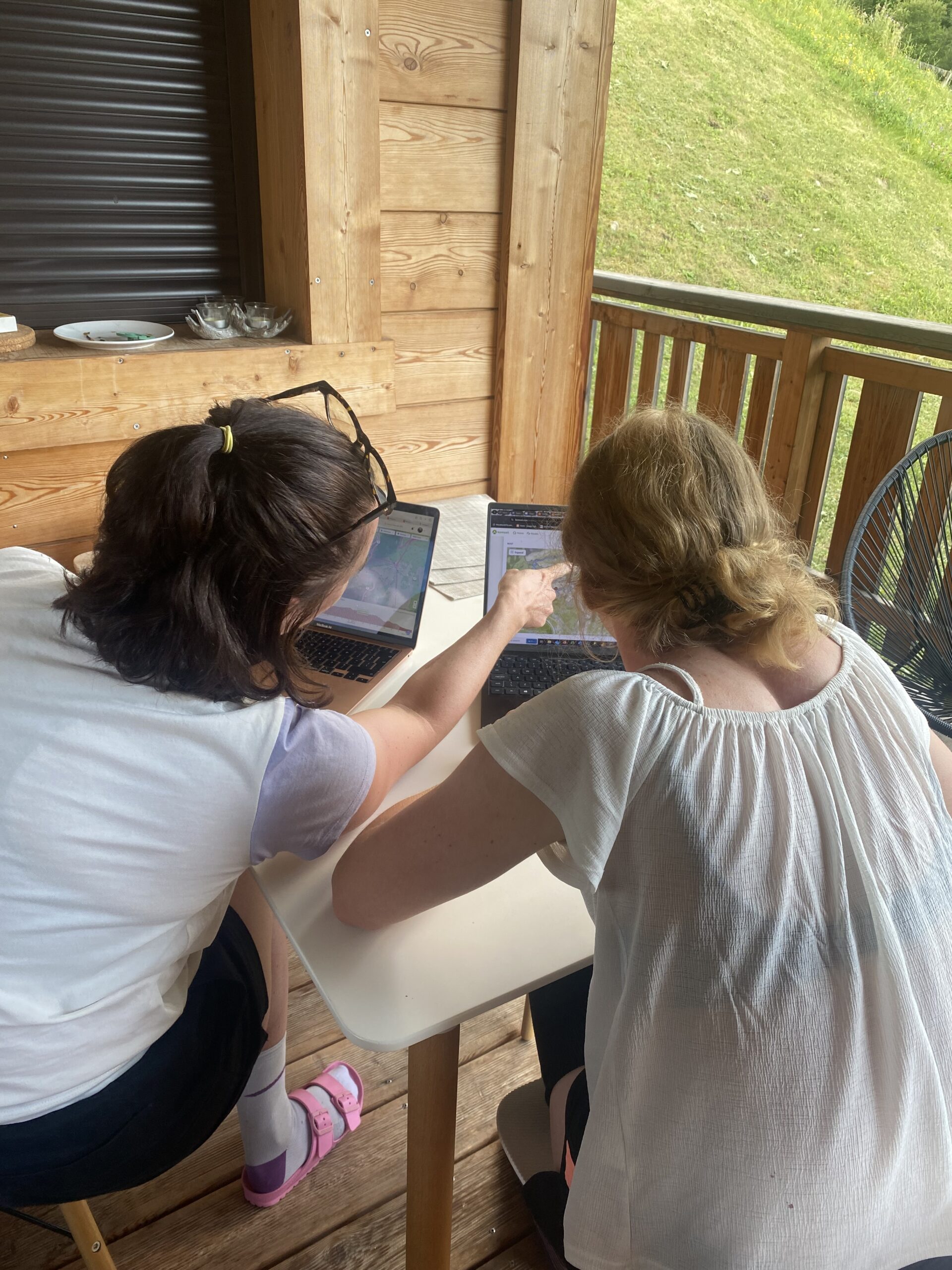 wo women sit on a wooden deck working on laptops, with one woman pointing at something on the other's screen. They are seated at a white table, focused on their work. The setting is outdoors, with a grassy hill in the background, and the deck features wooden walls and railings