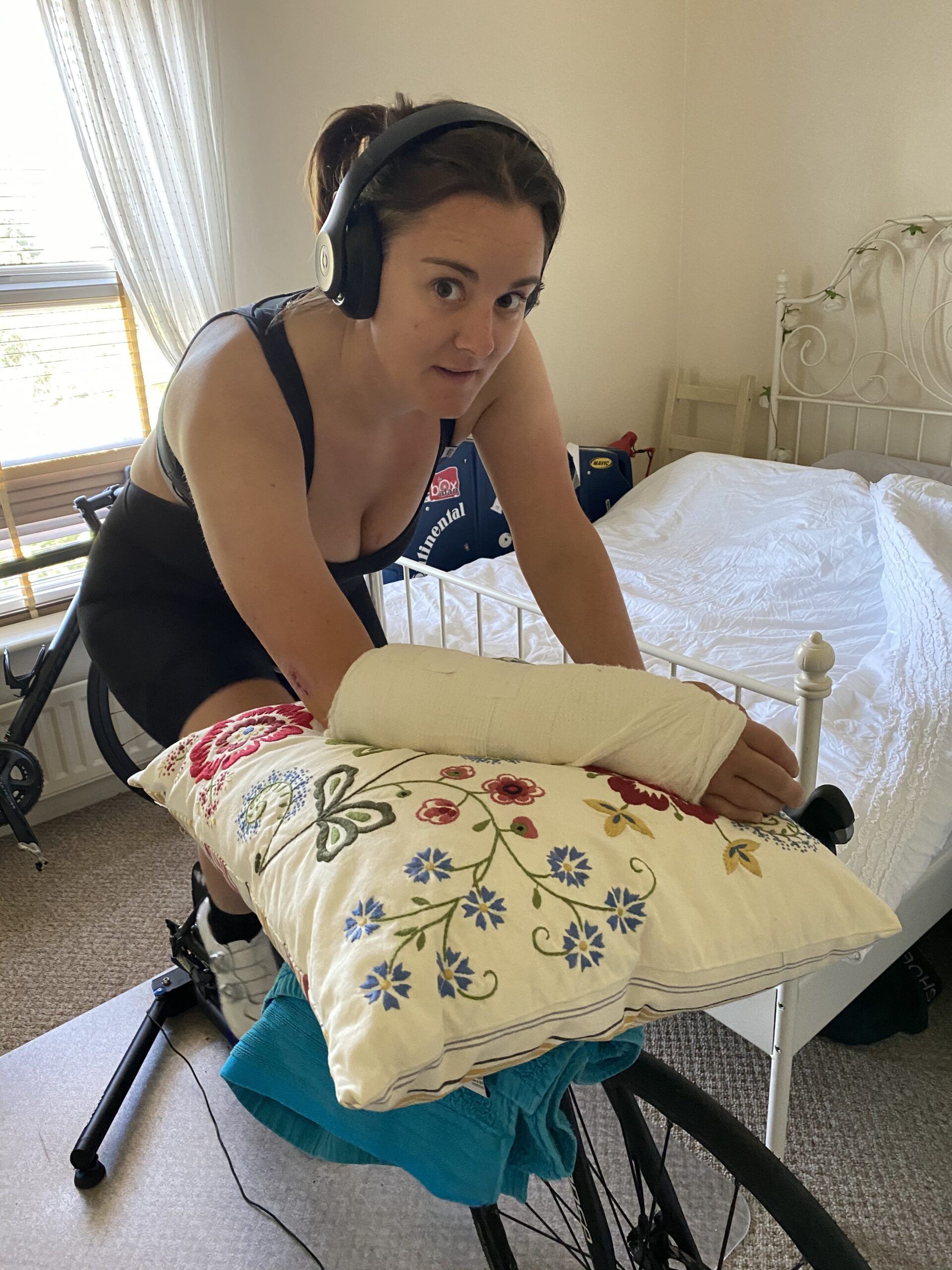 "A woman with headphones rides an indoor stationary bike in a bedroom. Her right arm is in a cast and resting on a floral pillow placed on the handlebars