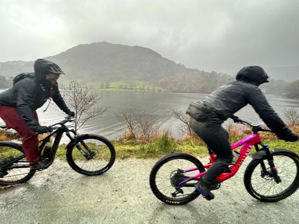 Two cyclists riding on a wet and windy day alongside a scenic lake and hills, wearing waterproof gear and helmets to stay warm and dry in challenging weather conditions