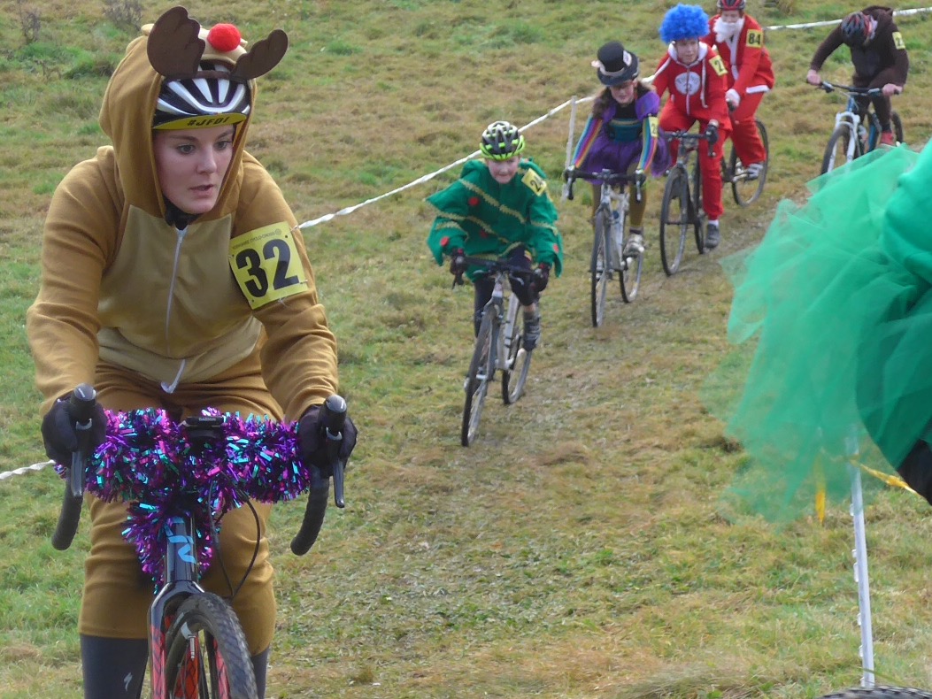 Festive cyclists in costumes, including a reindeer, elf, and Santa, riding in a grassy race adorned with colorful decorations and holiday cheer