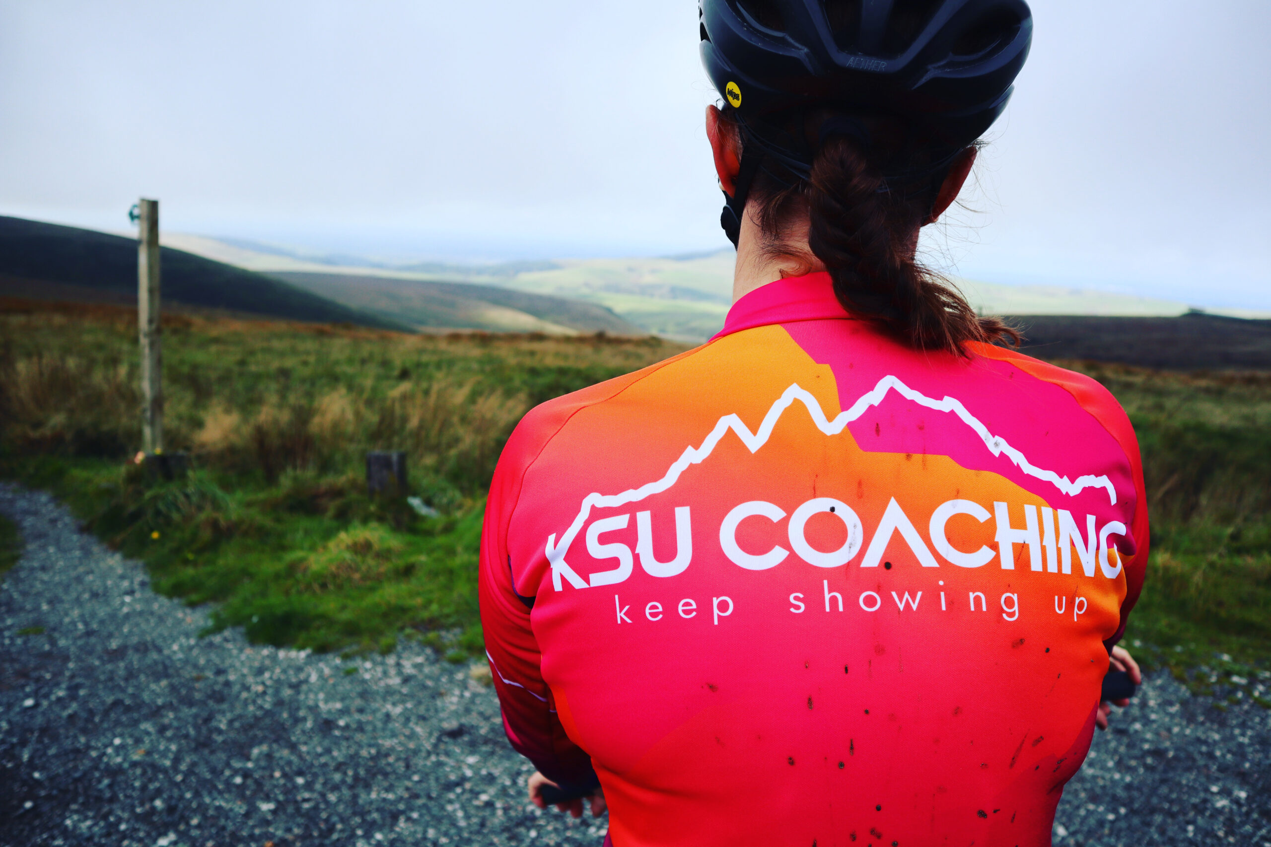 A cyclist wearing a pink and orange "KSU COACHING" jersey with "keep showing up" on the back, set against a scenic gravel path and misty hills.